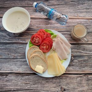 Zdjęcie przedstawia Dietę podstawową:  Kawa zbożowa z mlekiem Owsianka z jabłkami Chleb mieszany pszenno-żytni Masło extra Szynka tostowa- drobiowa, średnio rozdrob. Ser żółty Pomidor Woda źródlana Sałata zielona